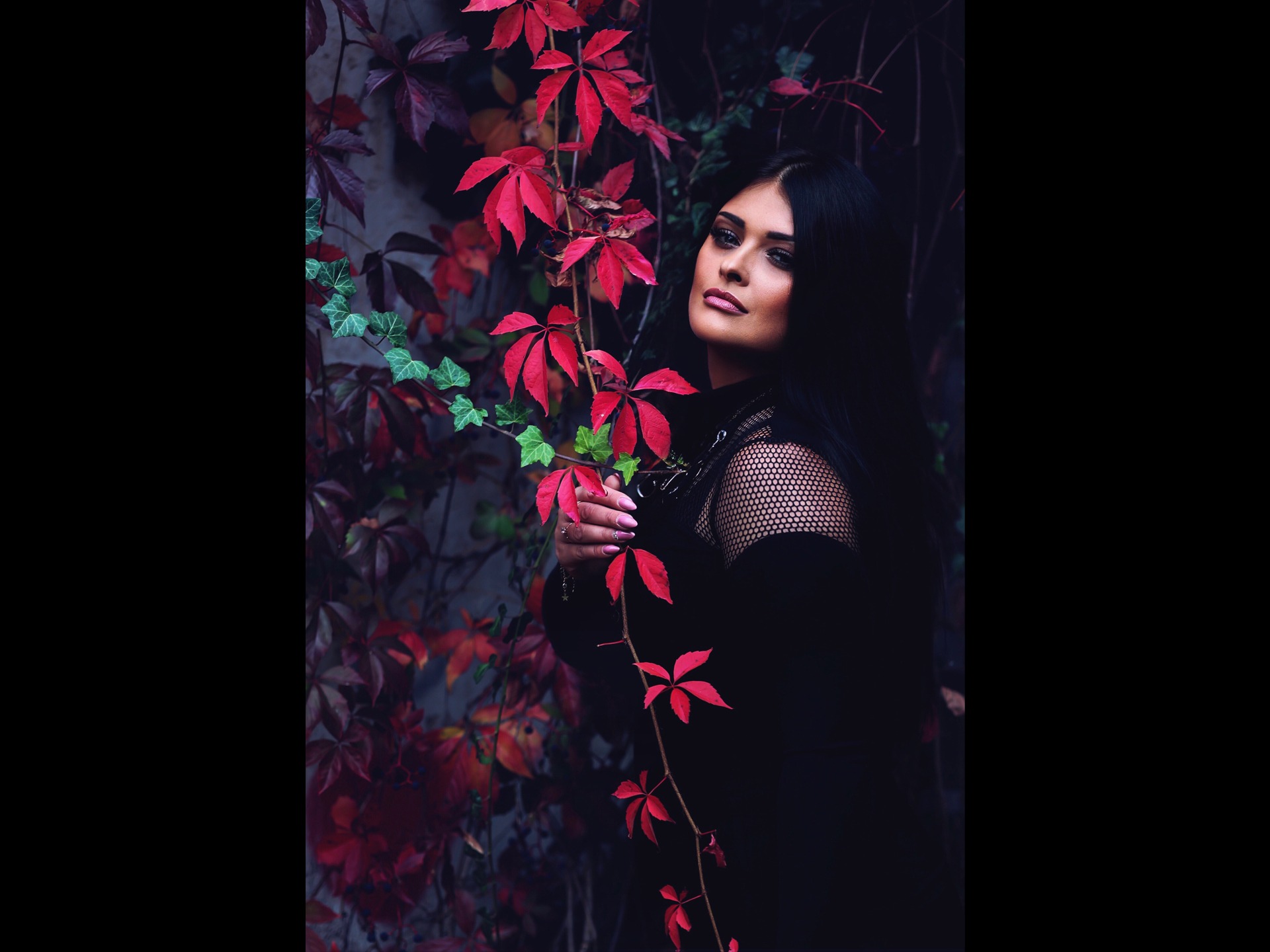 Portret kobiety z ciemnymi włosami, pozującej na tle ściany z czerwonymi i zielonymi liśćmi bluszczu.
