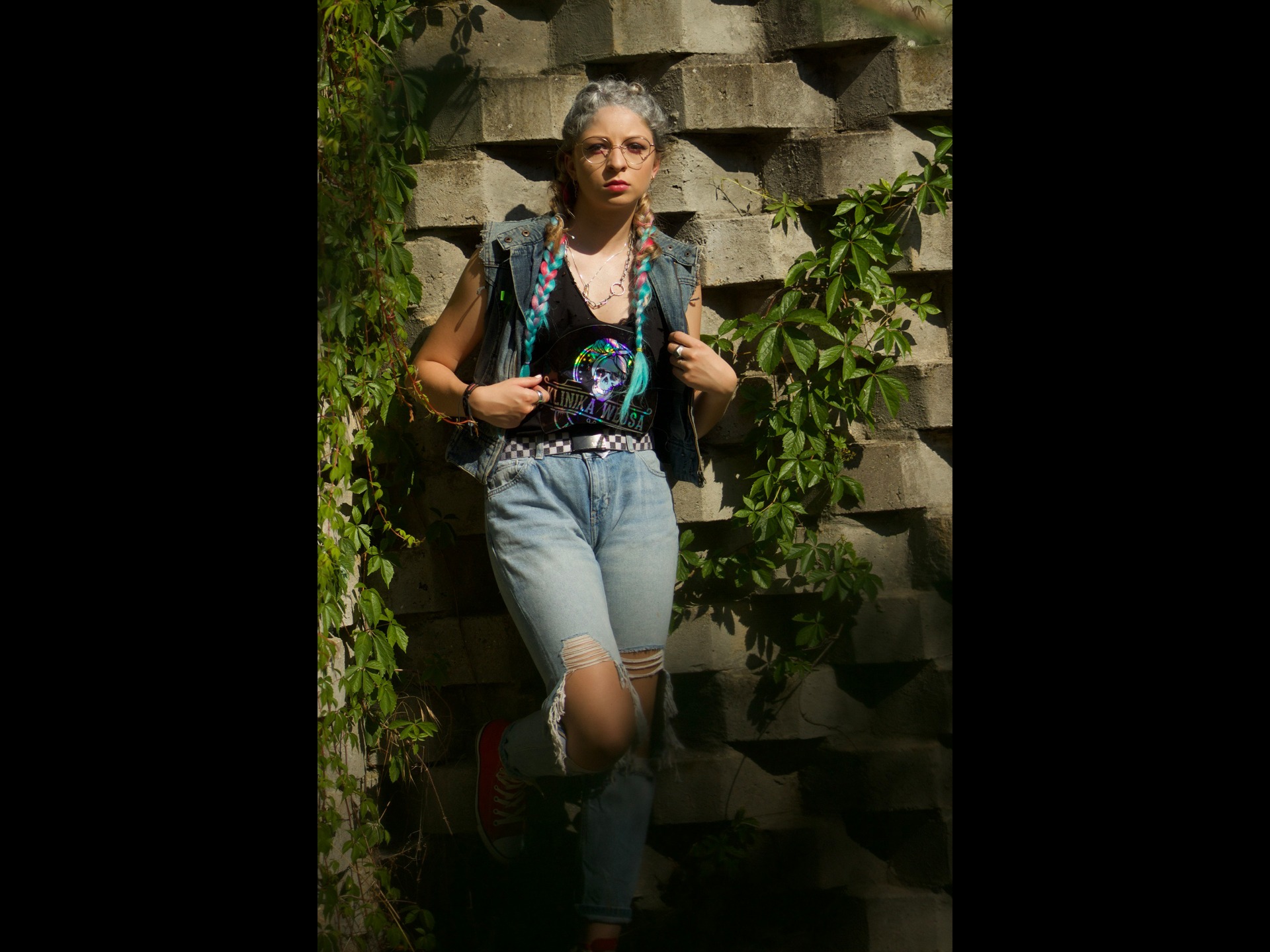 Młoda kobieta z kolorowymi warkoczami i okularami, ubrana w dżinsową kamizelkę i podkoszulek z nadrukiem, pozuje oparta o betonową ścianę porośniętą bluszczem.