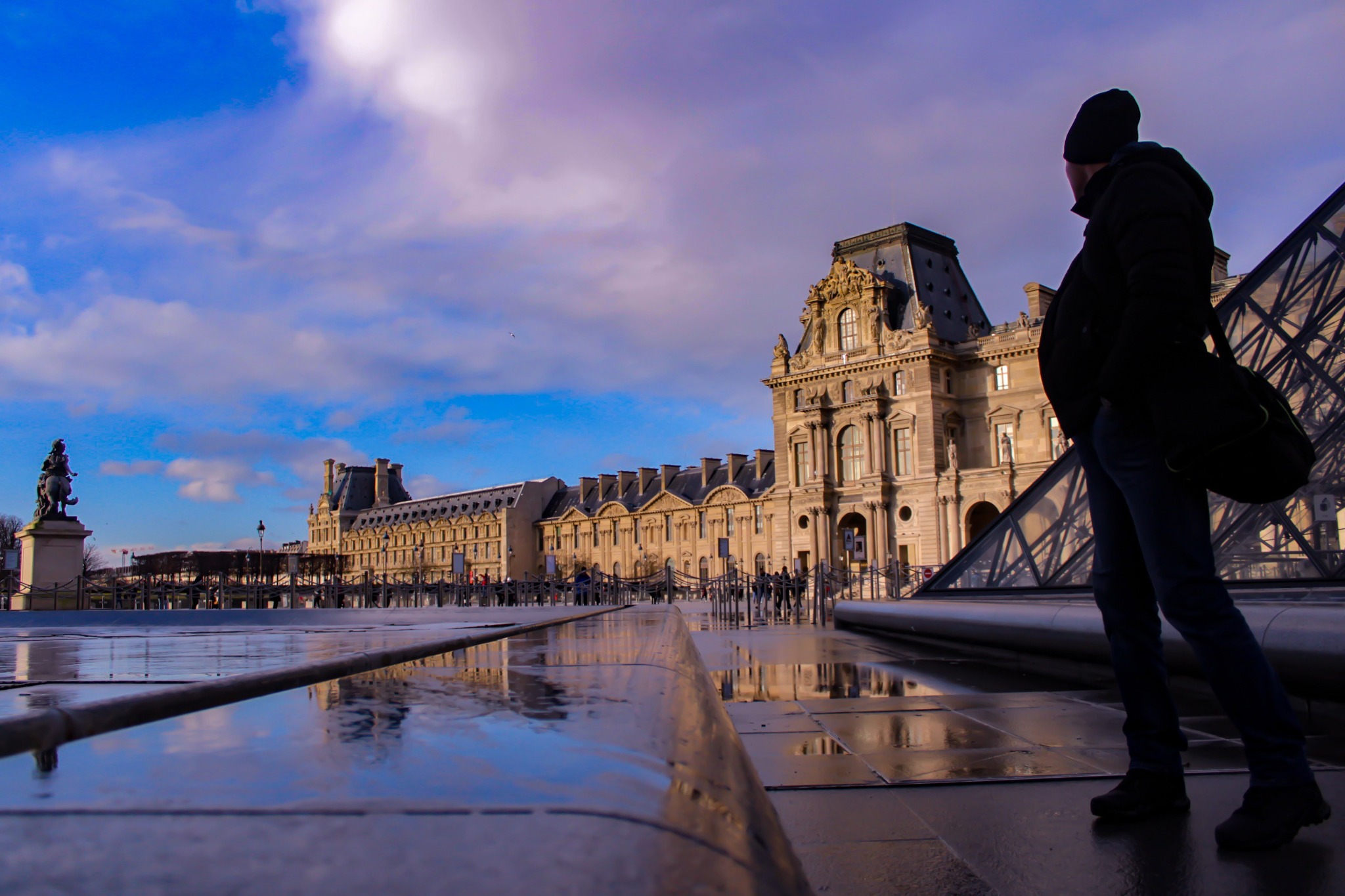 Mężczyzna w czapce i kurtce, z torbą na ramieniu, stoi na tle Luwru i jego piramidy, z odbiciem w mokrej nawierzchni.