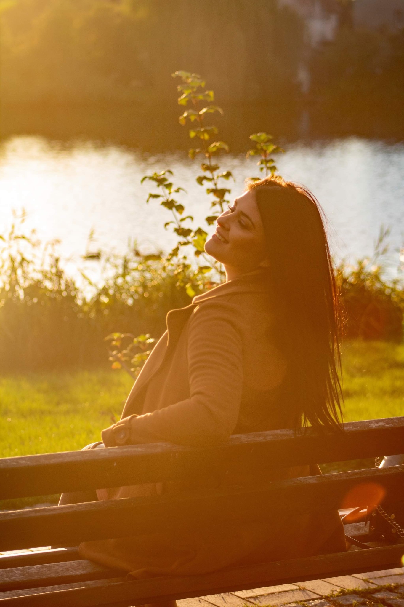 Portret kobiety w płaszczu siedzącej na ławce w parku, z widokiem na jezioro w złotym świetle zachodzącego słońca.