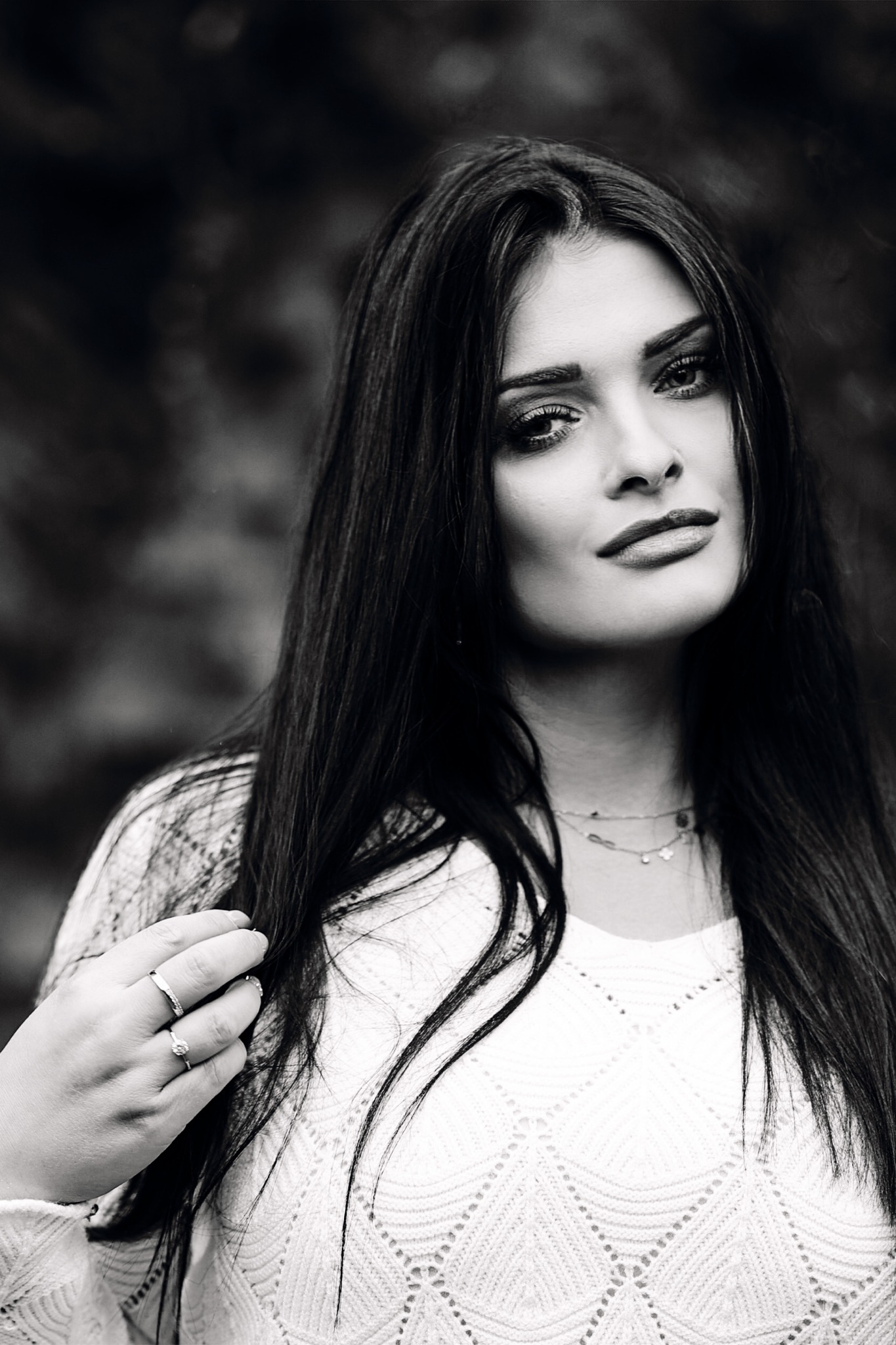 Portret młodej kobiety o długich, ciemnych włosach, ubranej w biały sweter o ażurowym wzorze, z delikatną biżuterią i pierścionkami, na rozmytym, ciemnym tle.