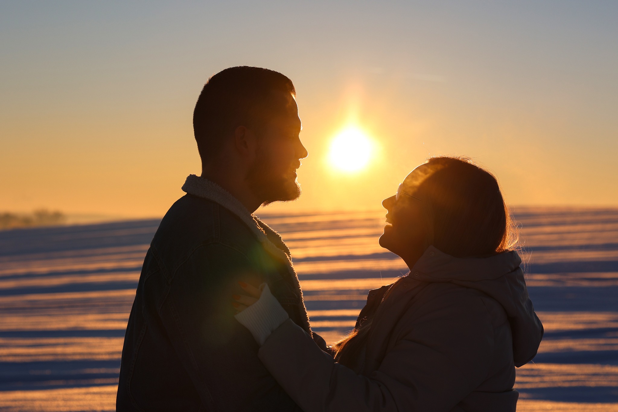 Zakochana para w zimowej scenerii, sylwetki na tle zachodzącego słońca, widoczne promienie słoneczne odbijające się od śniegu.