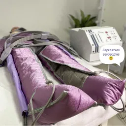 Klientka podczas zabiegu drenażu limfatycznego nóg w fioletowym kombinezonie, obok białe urządzenie sterujące i dekoracyjna roślina.