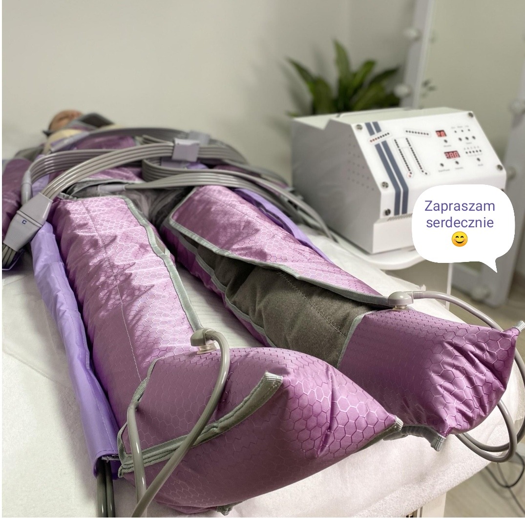 Klientka podczas zabiegu drenażu limfatycznego nóg w fioletowym kombinezonie, obok białe urządzenie sterujące i dekoracyjna roślina.