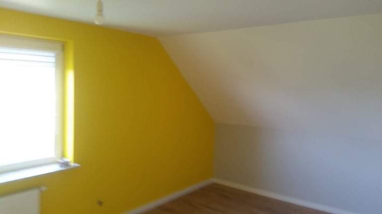 Pokój po malowaniu: jedna ściana w odcieniu żółci, druga w jasnym beżu, drewniana podłoga, białe okno z roletami, widoczny grzejnik.