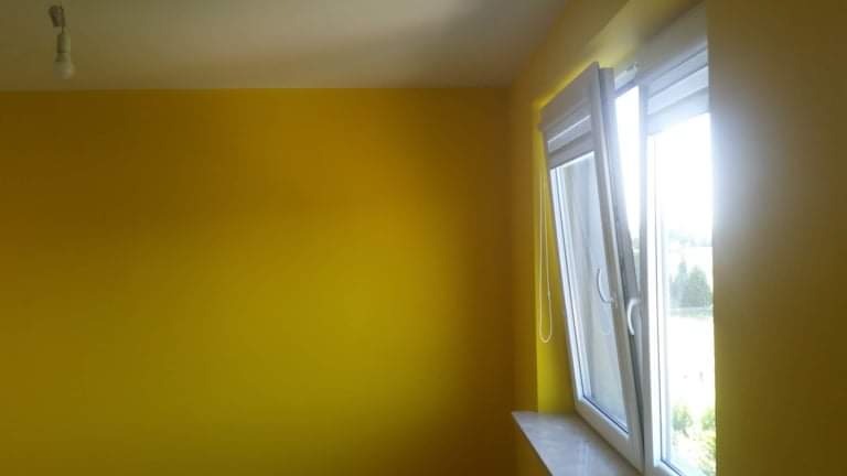 Ściana pomalowana na intensywny żółty kolor w pokoju z otwartym, białym oknem PCV, widok na zieleń za oknem.