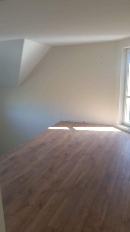 Wykończone poddasze z panelami podłogowymi w odcieniach brązu, widok na okno i skośną ścianę pomalowaną na biało, wpadające promienie słoneczne.