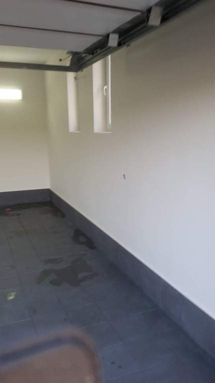 Wnętrze garażu po malowaniu ścian na biało, z ciemną podłogą z płytek i widocznymi dwoma oknami w górnej części ściany.