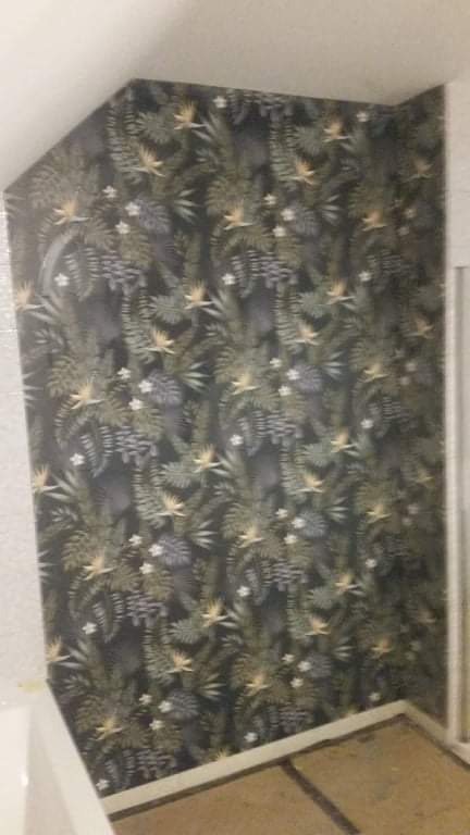 Ściana w rogu pokoju pokryta tapetą w tropikalne rośliny i kwiaty, widoczny fragment podłogi i listwy przypodłogowej.