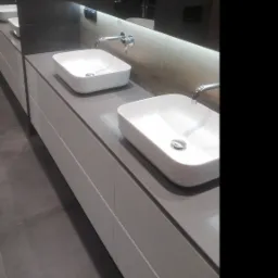 Dwie białe, prostokątne umywalki nablatowe z chromowanymi bateriami, zamontowane na szarym blacie z szufladami w nowoczesnej łazience z ciemnymi płytkami i podświetlanym lustrem.