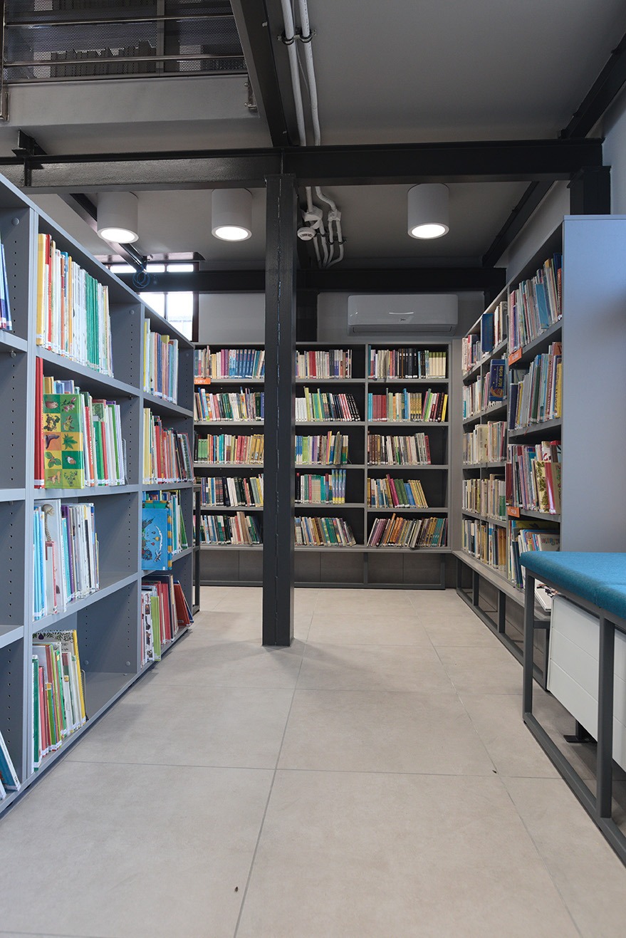 Wnętrze biblioteki z regałami wypełnionymi książkami, oświetlone lampami sufitowymi i z widoczną instalacją klimatyzacji.