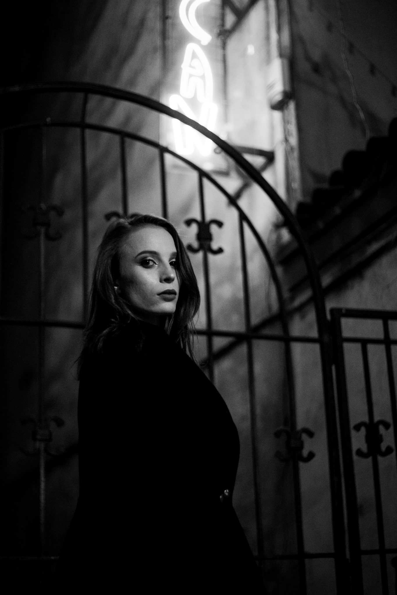 Portret kobiety w czarnym golfie na tle metalowej bramy i neonu w stylu retro, czarno-biała fotografia.