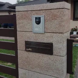 Szary wideodomofon z kamerą i brązowa skrzynka pocztowa zamontowane na słupku ogrodzeniowym z beżowego kamienia łupanego.