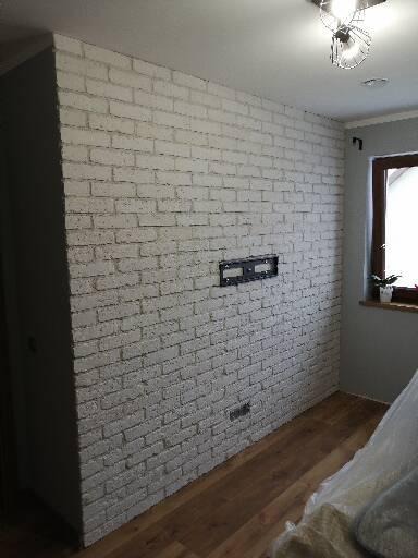 Ściana z białej cegły dekoracyjnej z przygotowanym miejscem na telewizor w Babimoście, widoczne drewniane panele podłogowe i okno z rośliną.