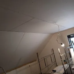 Poddasze z zamontowanymi płytami kartonowo-gipsowymi, widoczne rusztowanie i okno dachowe, instalacja elektryczna z żarówką tymczasowo zamontowana, ślady po szpachlowaniu na łączeniach płyt.