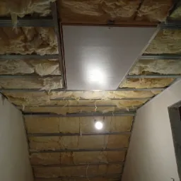 Niedokończone ocieplenie poddasza z widoczną wełną mineralną pomiędzy metalowymi profilami, częściowo zakryte płytą inspekcyjną i oświetlone pojedynczą żarówką.