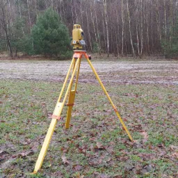 Żółty teodolit ustawiony na trójnogu na zewnątrz, na trawie pokrytej opadłymi liśćmi, z lasem w tle.