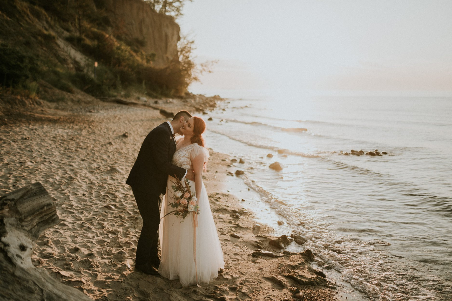 Para młoda w objęciach na plaży o zachodzie słońca, panna młoda z rudymi włosami trzyma bukiet, w tle klif i morze.