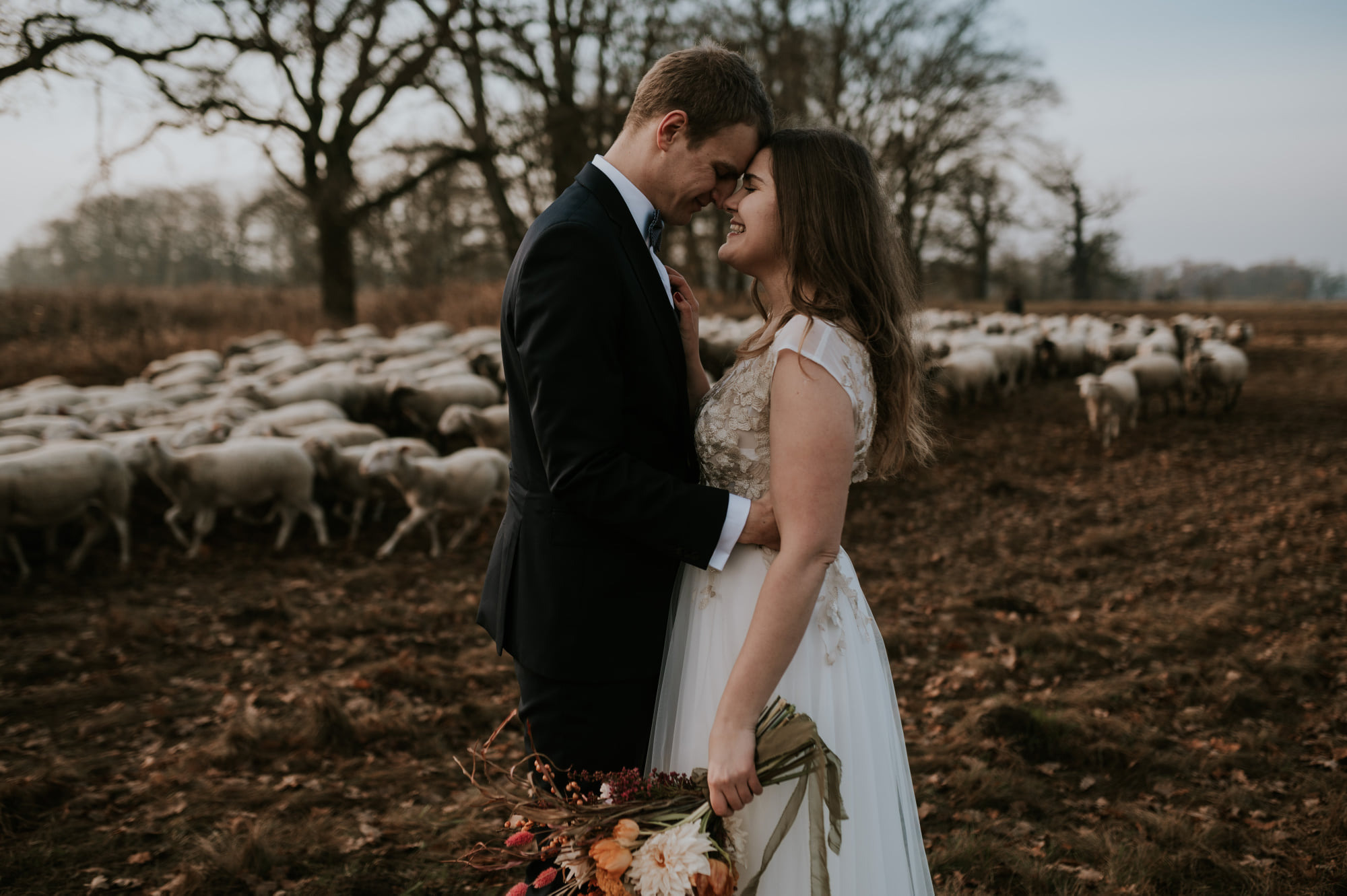 Para młoda przytulona na łące z pasącymi się owcami w tle, ona w sukni z koronkową górą i bukietem, on w czarnym garniturze.