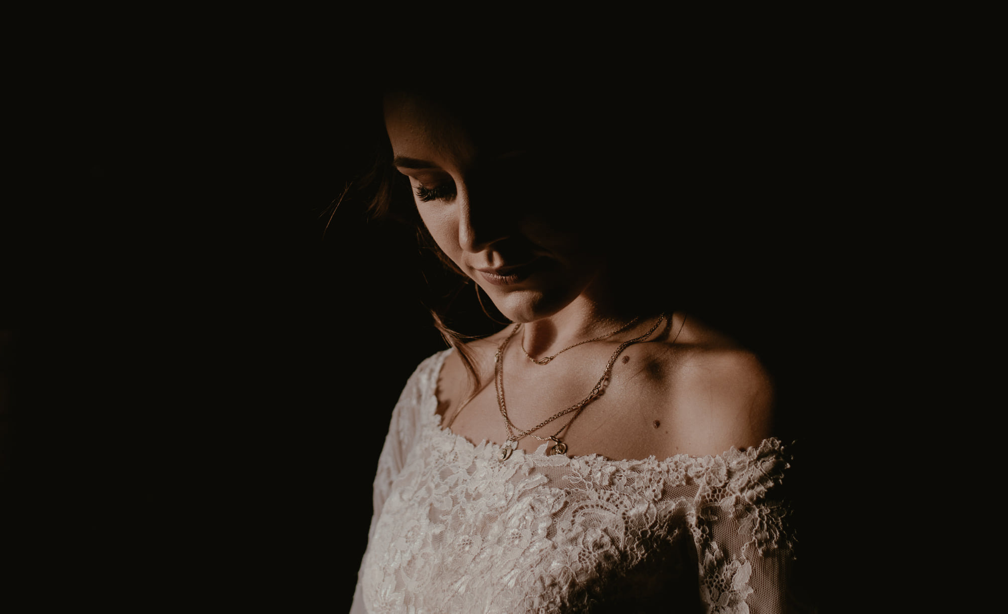 Portret panny młodej w koronkowej sukni z dekoltem carmen i złotym łańcuszkiem, oświetlona z góry, z twarzą częściowo w cieniu.