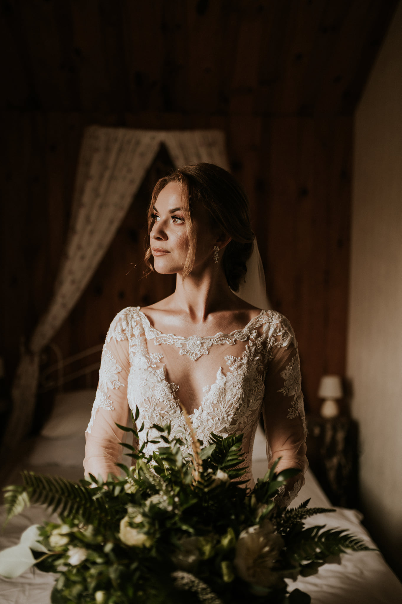 Panna młoda w koronkowej sukni z bukietem, spoglądająca w bok na tle drewnianej ściany i łóżka z baldachimem.