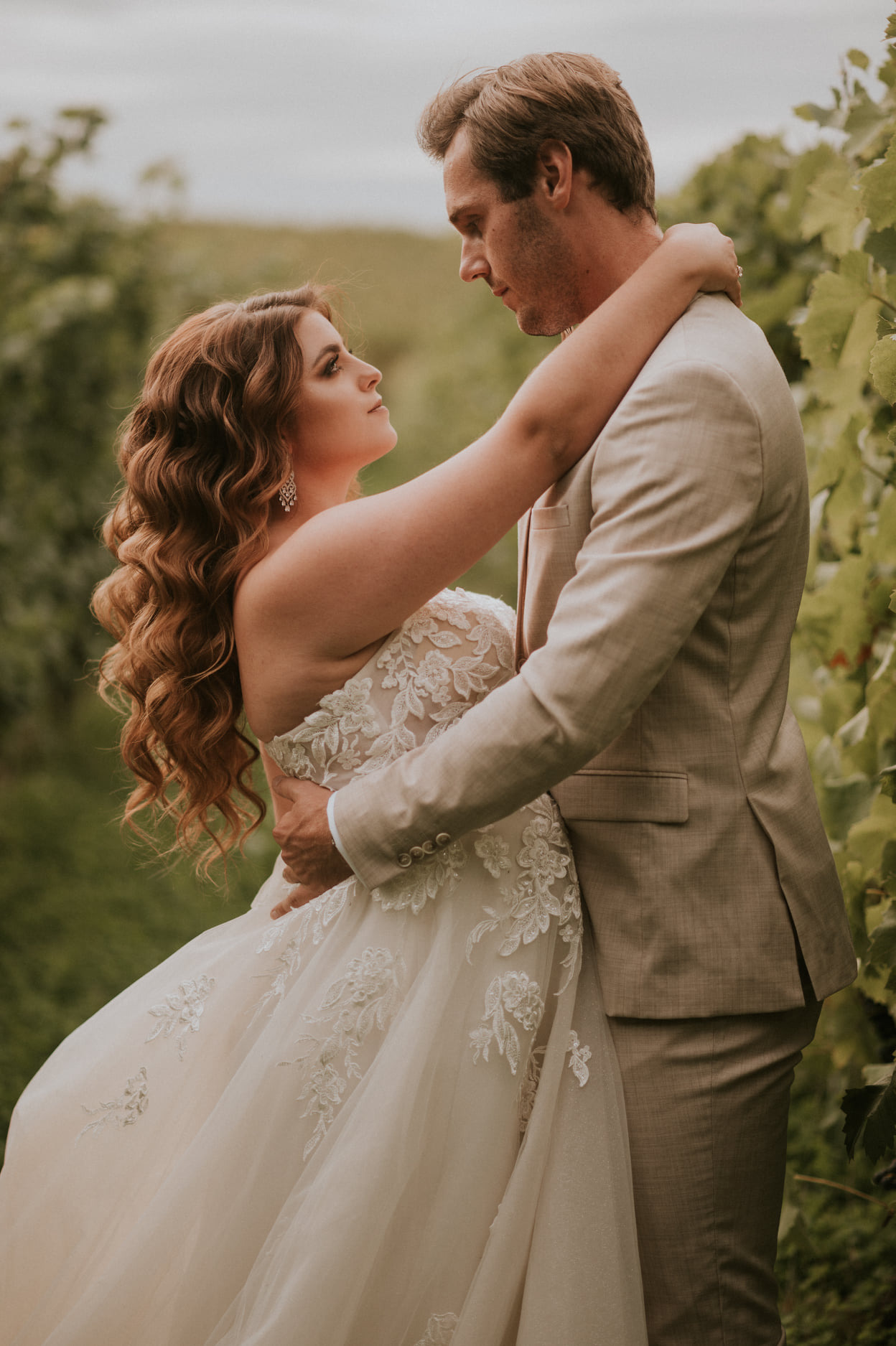 Para młoda w objęciach na tle winnicy, panna młoda w sukni z koronkowymi aplikacjami spogląda na pana młodego w beżowym garniturze.