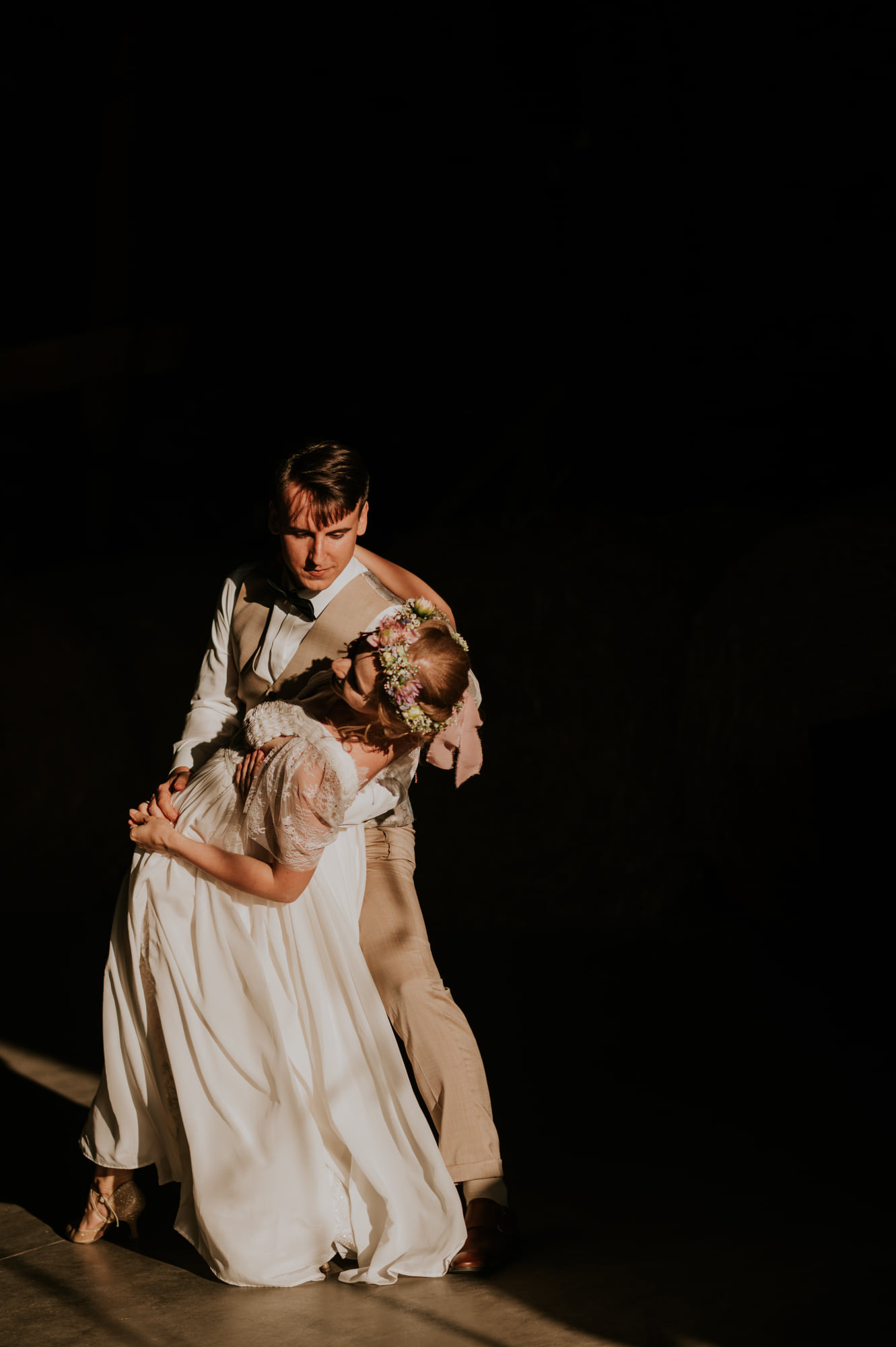 Para młoda w tańcu, ona w koronkowej sukni i wianku, on w beżowym garniturze, oświetleni pojedynczym promieniem światła na ciemnym tle.