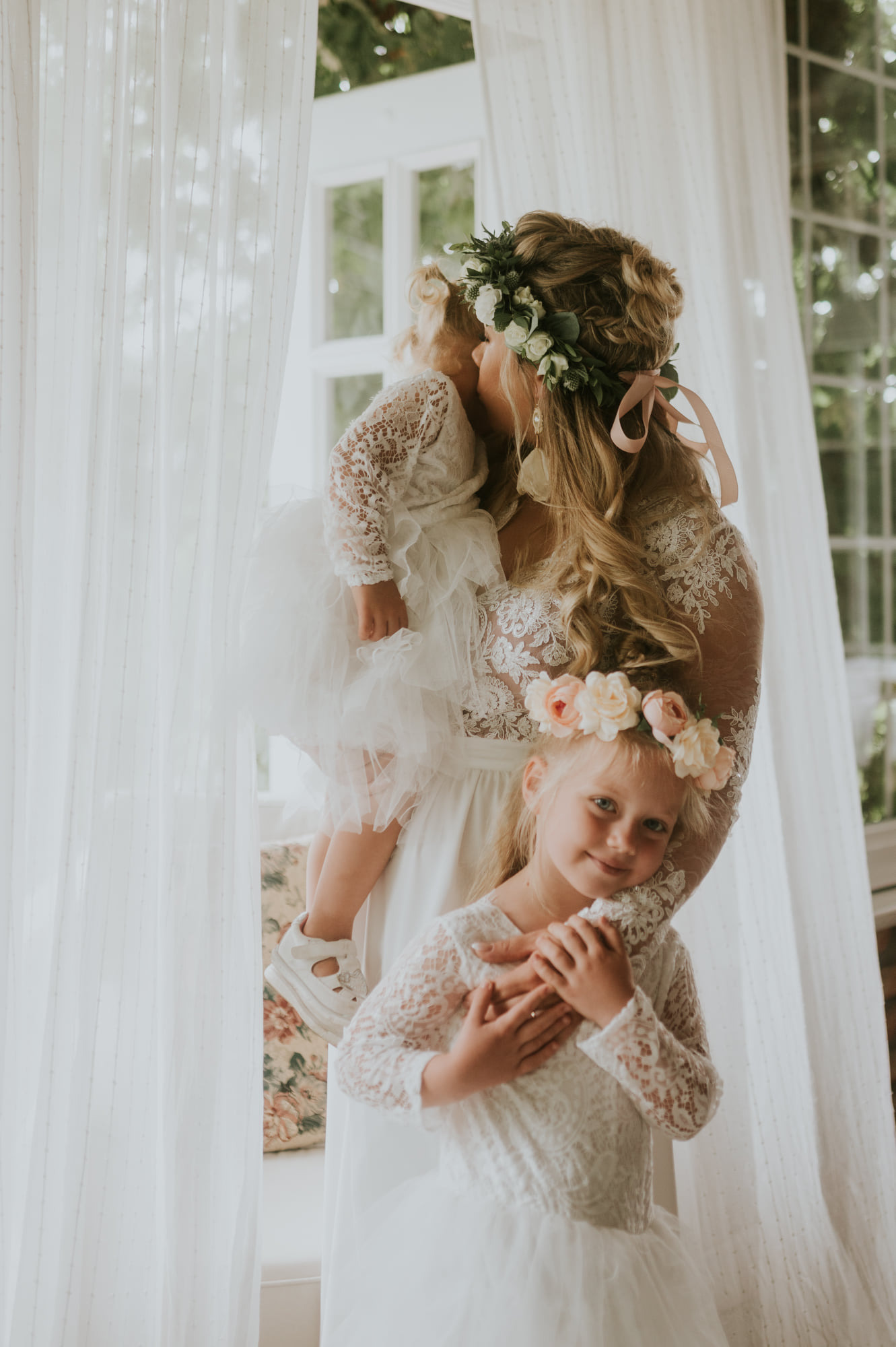 Mama w koronkowej sukni trzymająca na rękach córeczkę, druga córka opiera się o mamę, wszystkie ubrane w białe, koronkowe stroje i kwiatowe wianki, jasne tło okna z białymi zasłonami.