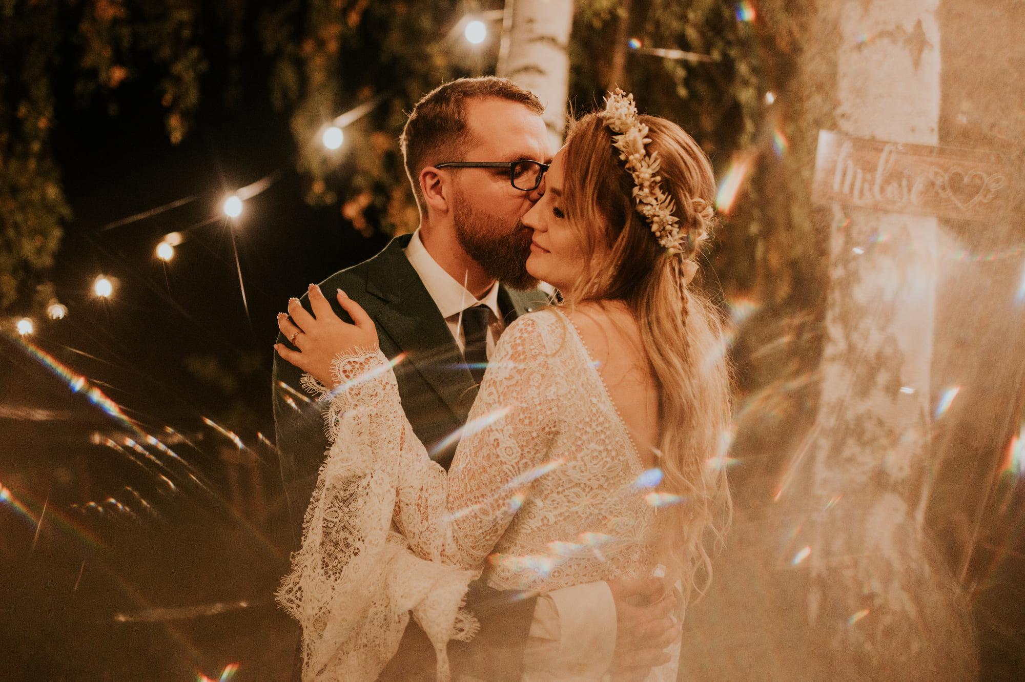 Panna młoda w koronkowej sukni i wianku obejmuje pana młodego w ciemnozielonym garniturze na tle drzewa z napisem 'Miłość' i girlandy światełek, z efektem rozproszonego światła.