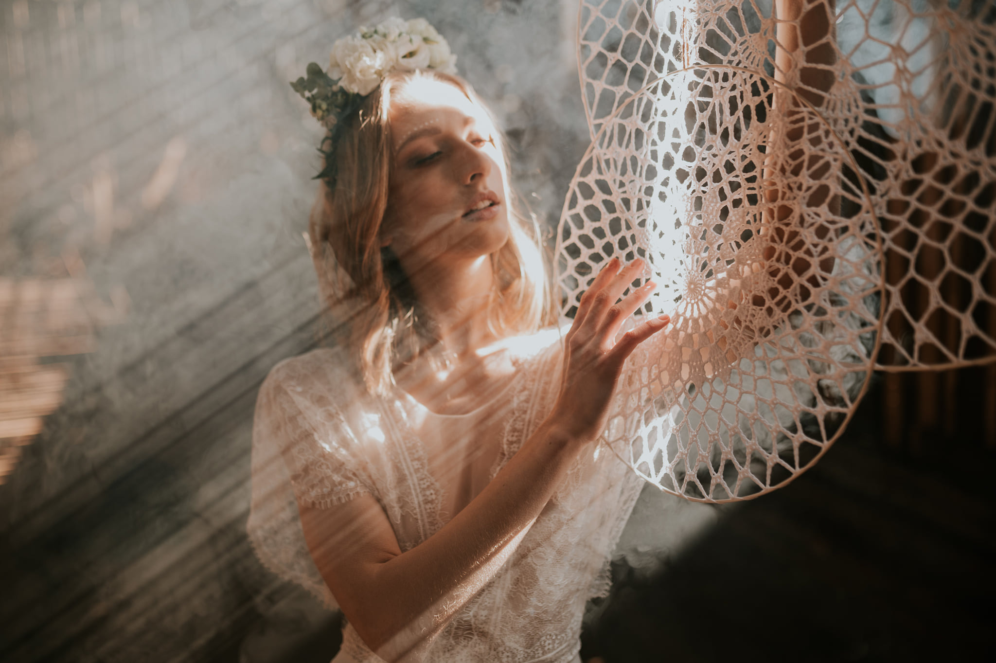 Portret kobiety w koronkowej sukience i wianku na głowie, delikatnie dotykającej ręką koronkowego łapacza snów w miękkim, rozproszonym świetle.
