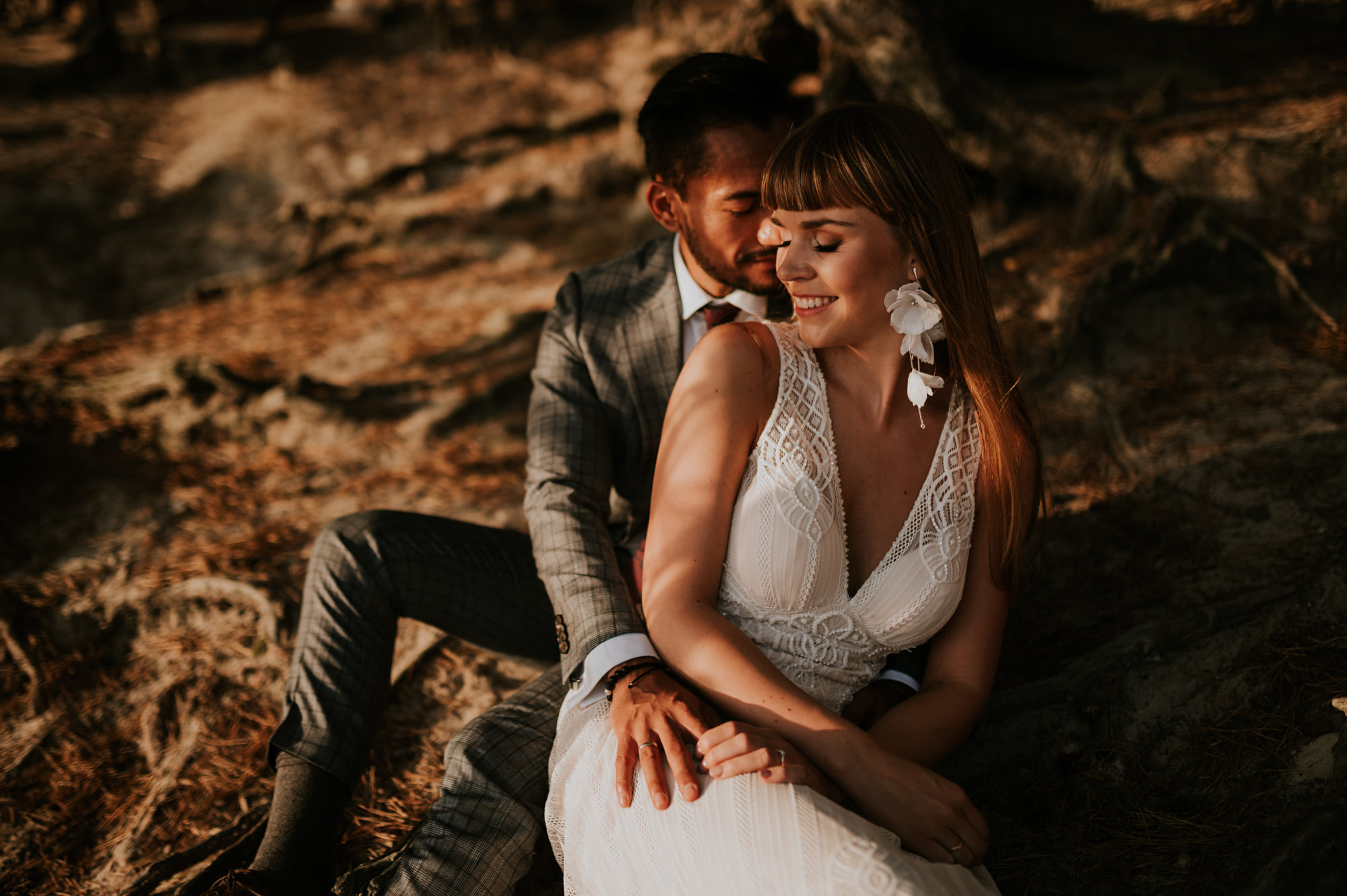 Uśmiechnięta panna młoda w koronkowej sukni z kwiatowymi kolczykami, obejmowana przez pana młodego w kraciastej marynarce na tle ziemi.