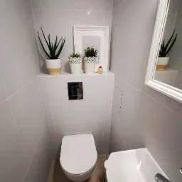 Nowoczesna, minimalistyczna łazienka w szarościach z wiszącą toaletą, umywalką narożną i dekoracyjnymi roślinami w doniczkach na półce, oświetlenie punktowe.