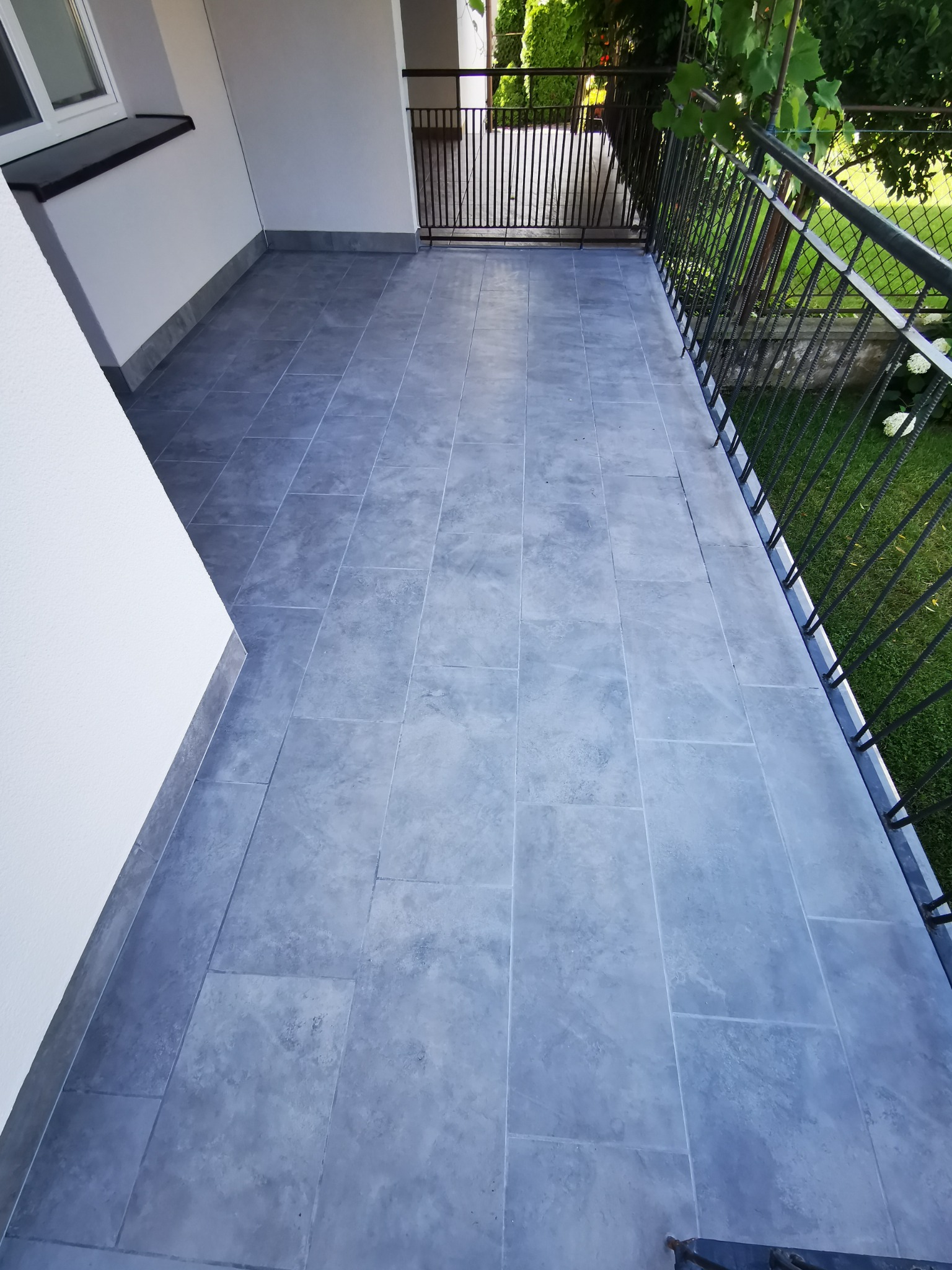 Wyłożony szarymi płytkami taras z czarną metalową balustradą, widok na zielony ogród.