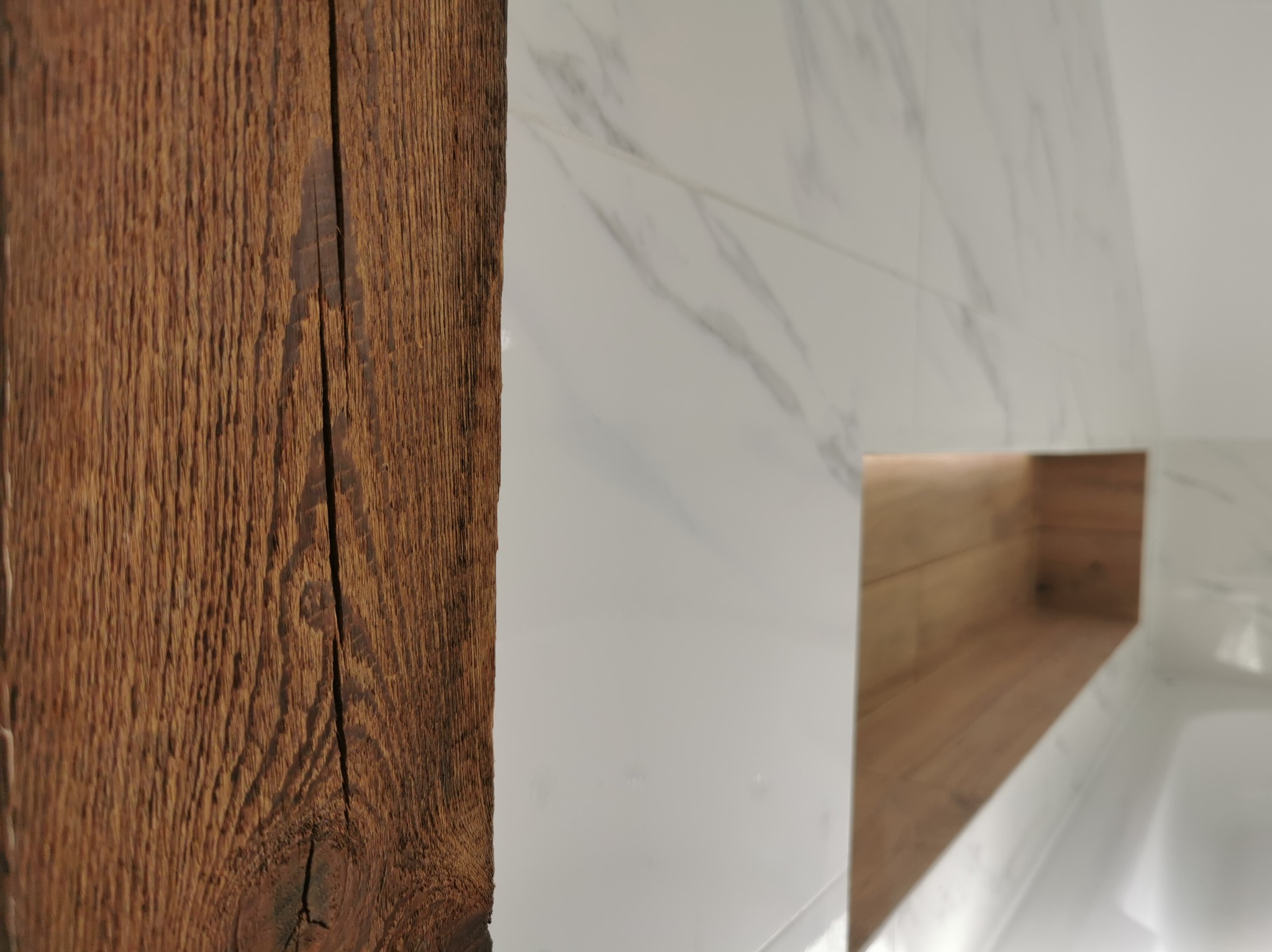 Fragment łazienki z białą marmurową ścianą, drewnianą półką wnękową z podświetleniem i elementem starego, ciemnego drewna z wyraźnym usłojeniem na pierwszym planie.