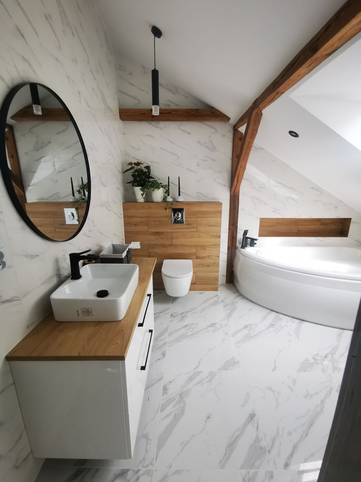 Nowoczesna łazienka na poddaszu z marmurowymi płytkami, drewnianymi belkami stropowymi, okrągłym lustrem w czarnej ramie, białą umywalką nablatową z czarną armaturą, wiszącą toaletą i okrągłą wanną.