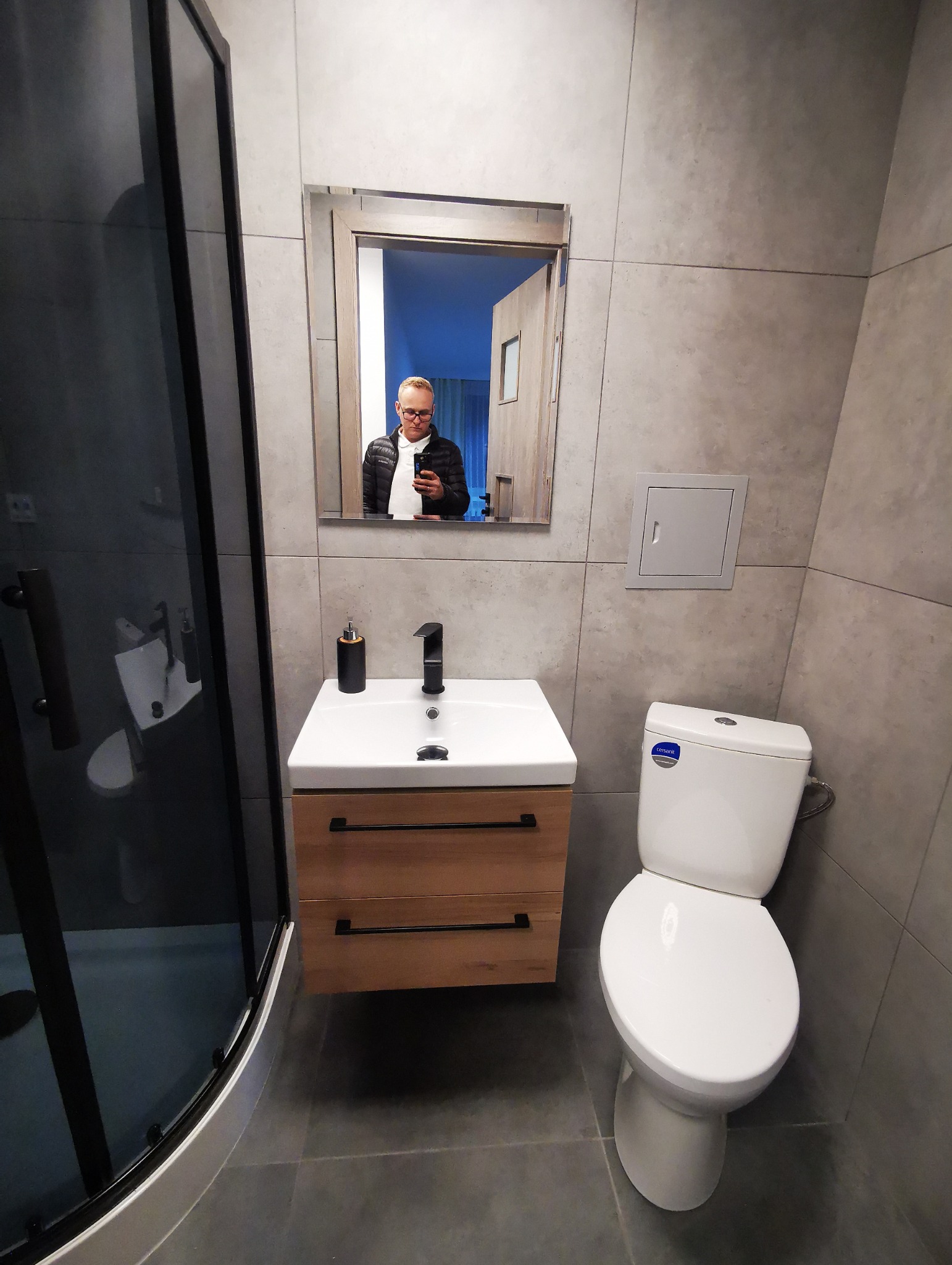 Nowoczesna łazienka z szarą glazurą, kabiną prysznicową z czarnym profilem, szafką pod umywalką z drewnianymi frontami i czarną armaturą, toaleta oraz odbicie osoby robiącej zdjęcie w lustrze.