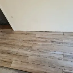 Ułożone płytki podłogowe imitujące drewno w pomieszczeniu, widoczna biała ściana z niedokończonym wykończeniem przy podłodze oraz wejście do innego pomieszczenia zabezpieczone folią.