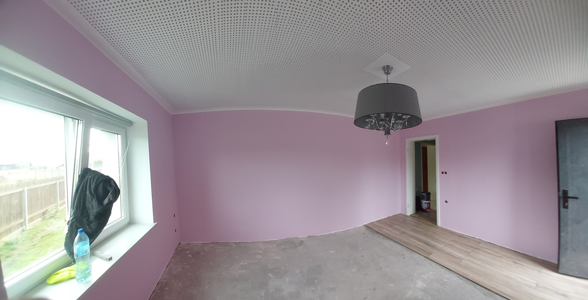 Pokój w trakcie prac wykończeniowych: ściany pomalowane na różowo, sufit z perforacją, częściowo ułożona podłoga z paneli, butelka wody i kurtka na parapecie.