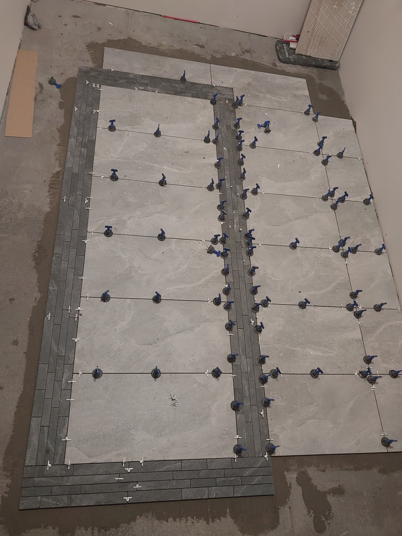 Układanie szarych płytek podłogowych z dekoracyjnym obramowaniem z ciemniejszych płytek, z użyciem systemu poziomowania z niebieskimi klipsami i krzyżykami dystansowymi.