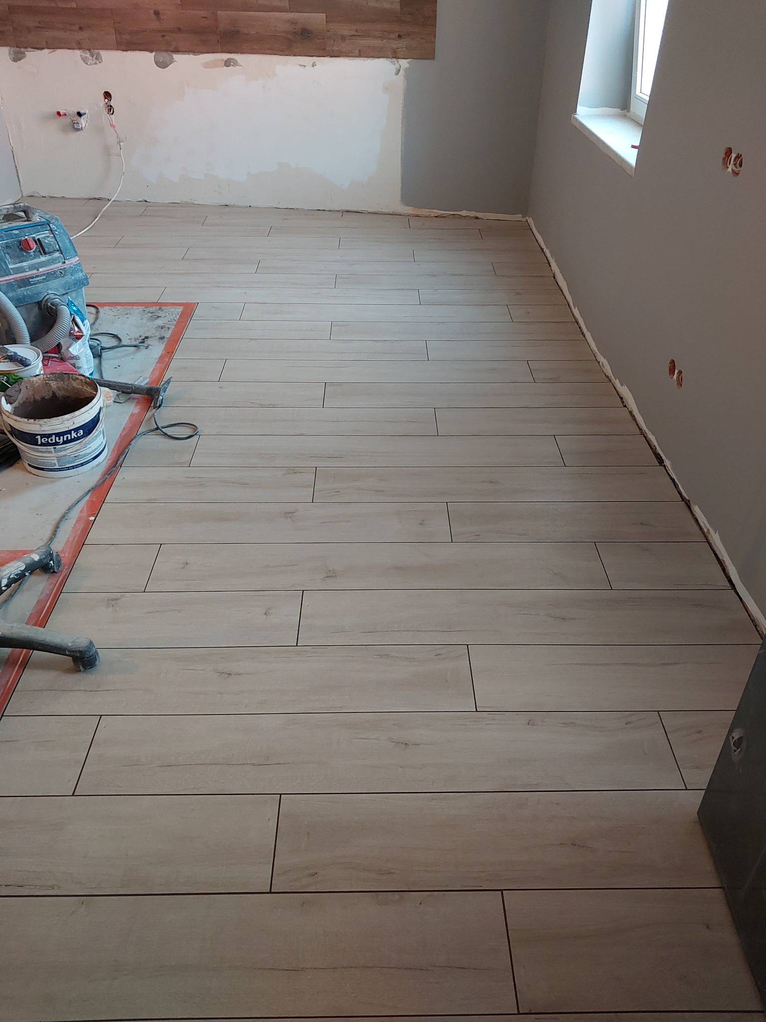 Ułożona podłoga z płytek imitujących drewno w trakcie remontu, widoczne narzędzia i materiały budowlane, ściany w szarym kolorze, brak wykończenia listew przypodłogowych.