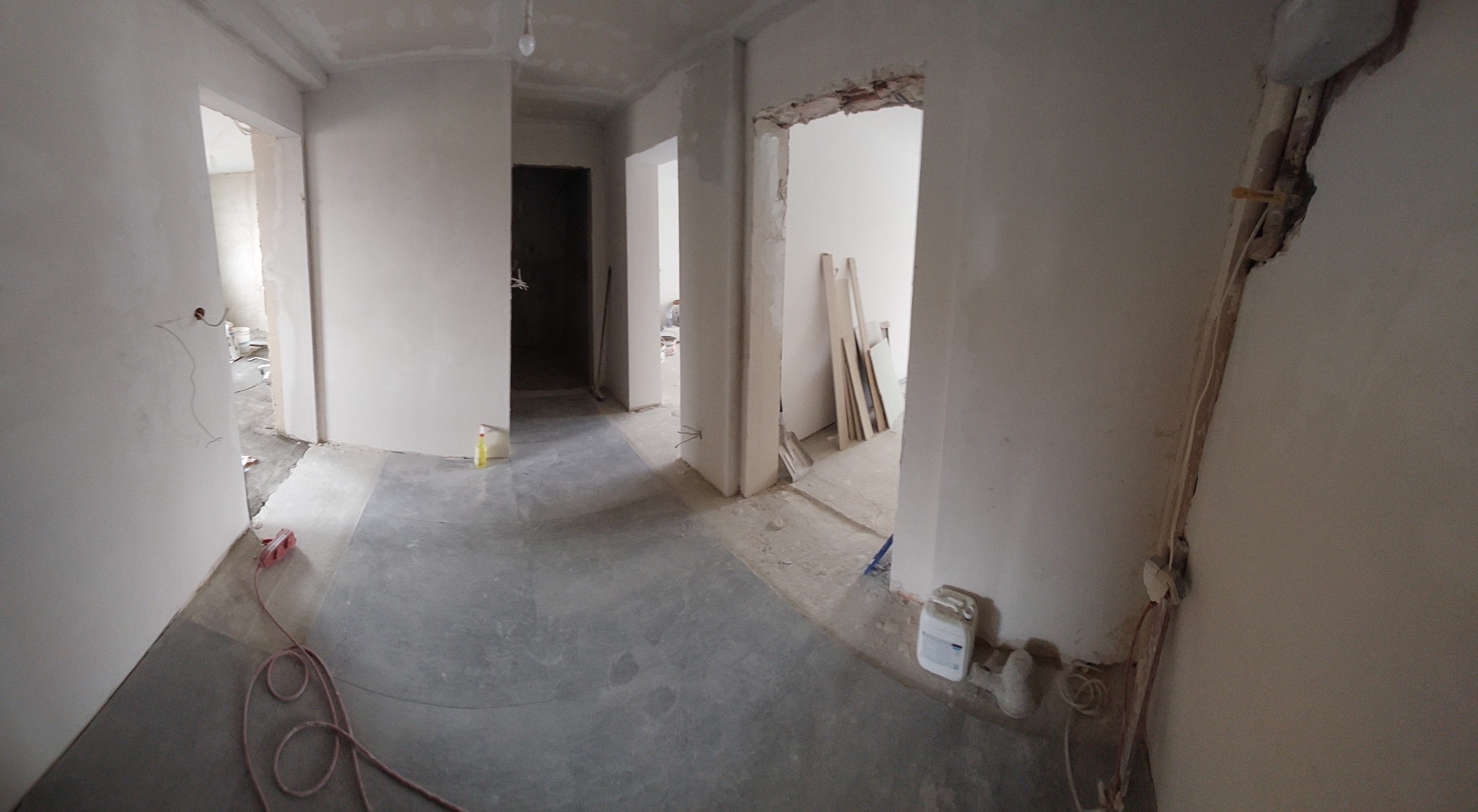 Wnętrze mieszkania w trakcie prac wykończeniowych: ściany po szpachlowaniu, przygotowane do dalszych prac, widoczne przewody elektryczne, przedłużacz i materiały budowlane.