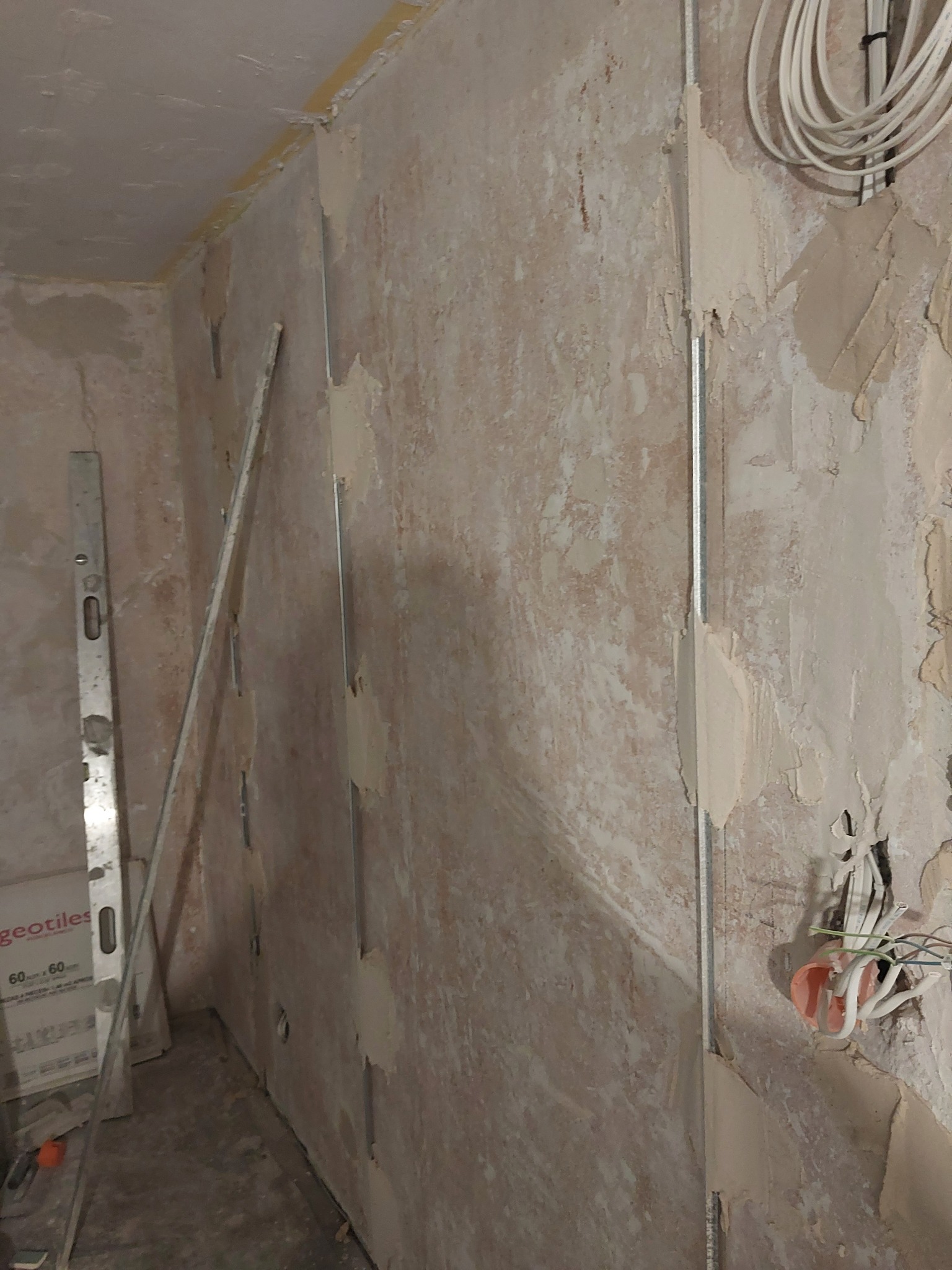 Przygotowanie ścian do wykończenia: metalowe profile pionowe przymocowane do nierównej ściany z pozostałościami starej farby i zaprawy, widoczna poziomica oparta o ścianę, przewody elektryczne...