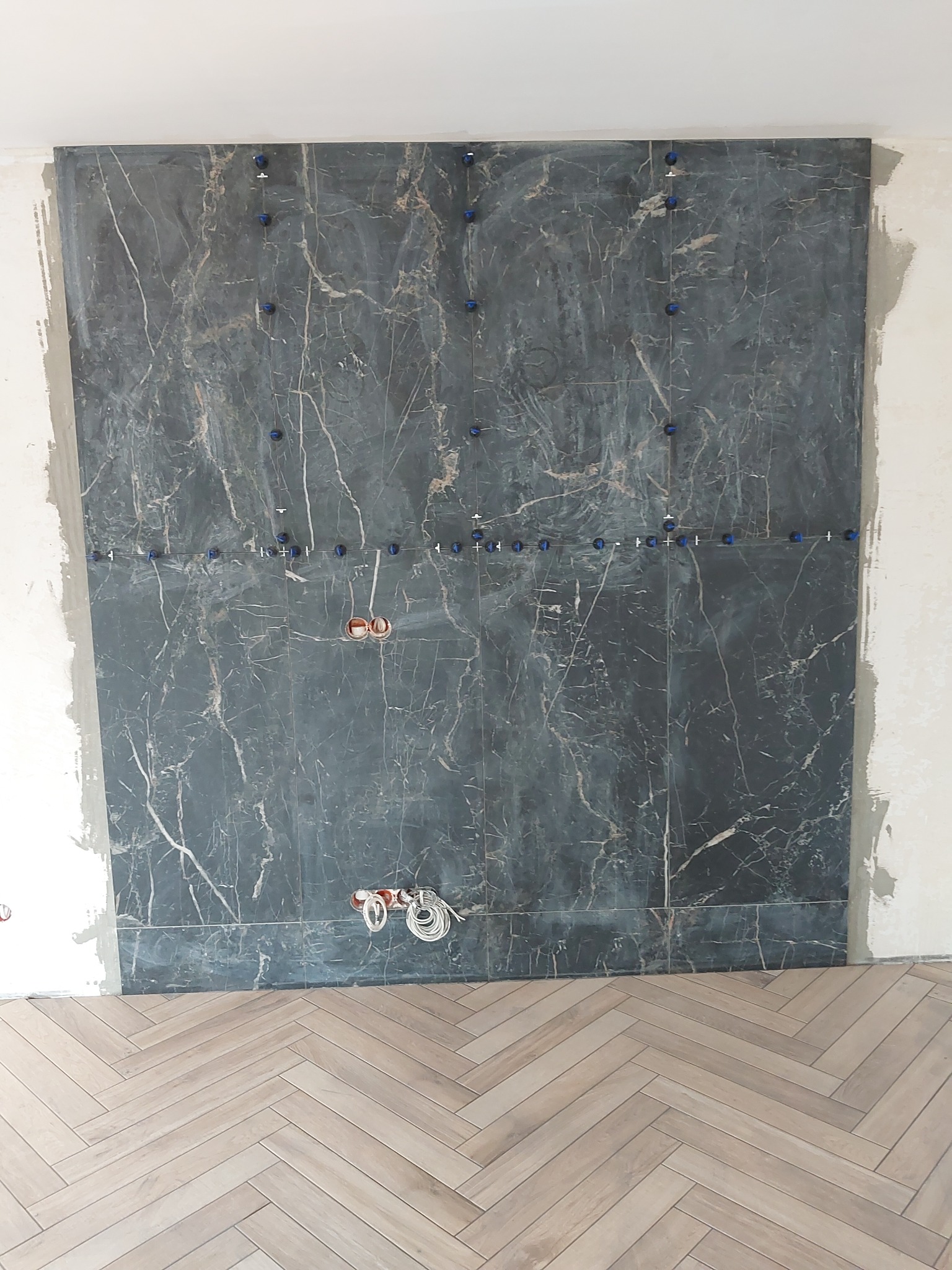 Ściana wyłożona wielkoformatowymi płytkami imitującymi marmur w trakcie montażu, widoczne niebieskie klipsy poziomujące i przewody elektryczne wystające ze ściany, na podłodze ułożony parkiet...