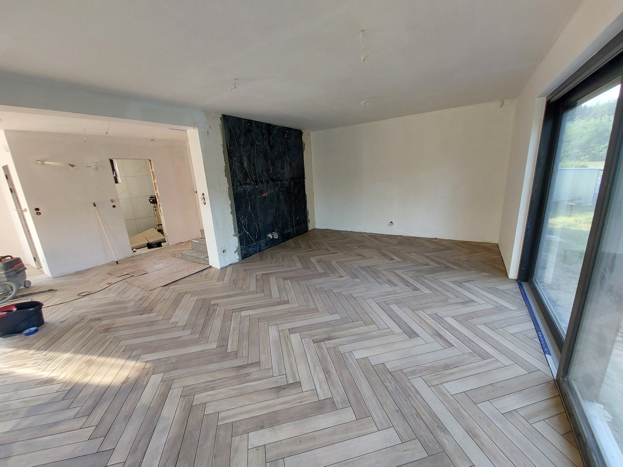 Jasna, drewnopodobna podłoga ułożona w jodełkę w przestronnym, częściowo wykończonym wnętrzu z dużym oknem i fragmentem ściany wyłożonym czarnym kamieniem.