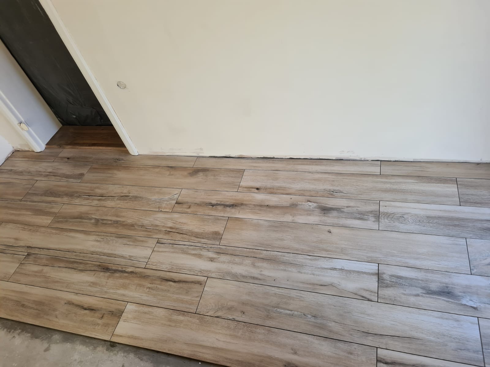 Ułożone płytki podłogowe imitujące drewno w pomieszczeniu, widoczna biała ściana z niedokończonym wykończeniem przy podłodze oraz wejście do innego pomieszczenia zabezpieczone folią.