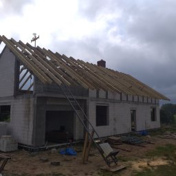 JAN BUD - Surowy stan budynku z pustaków z drewnianą konstrukcją dachu w trakcie budowy, widoczne narzędzia i materiały budowlane.