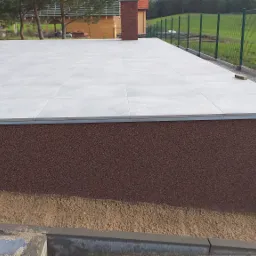 Jasnoszara, kwadratowa podłoga wyłożona płytkami na zewnątrz budynku, z widoczną elewacją w kolorze brązowym i elementami krajobrazu w tle.