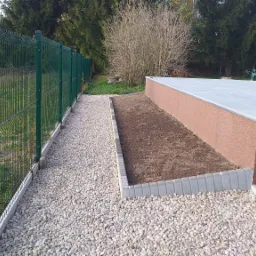 Wykończony taras z ceglanym kominem, obok rabata ogrodowa obłożona kostką i ścieżka z białego żwiru wzdłuż zielonego ogrodzenia panelowego.