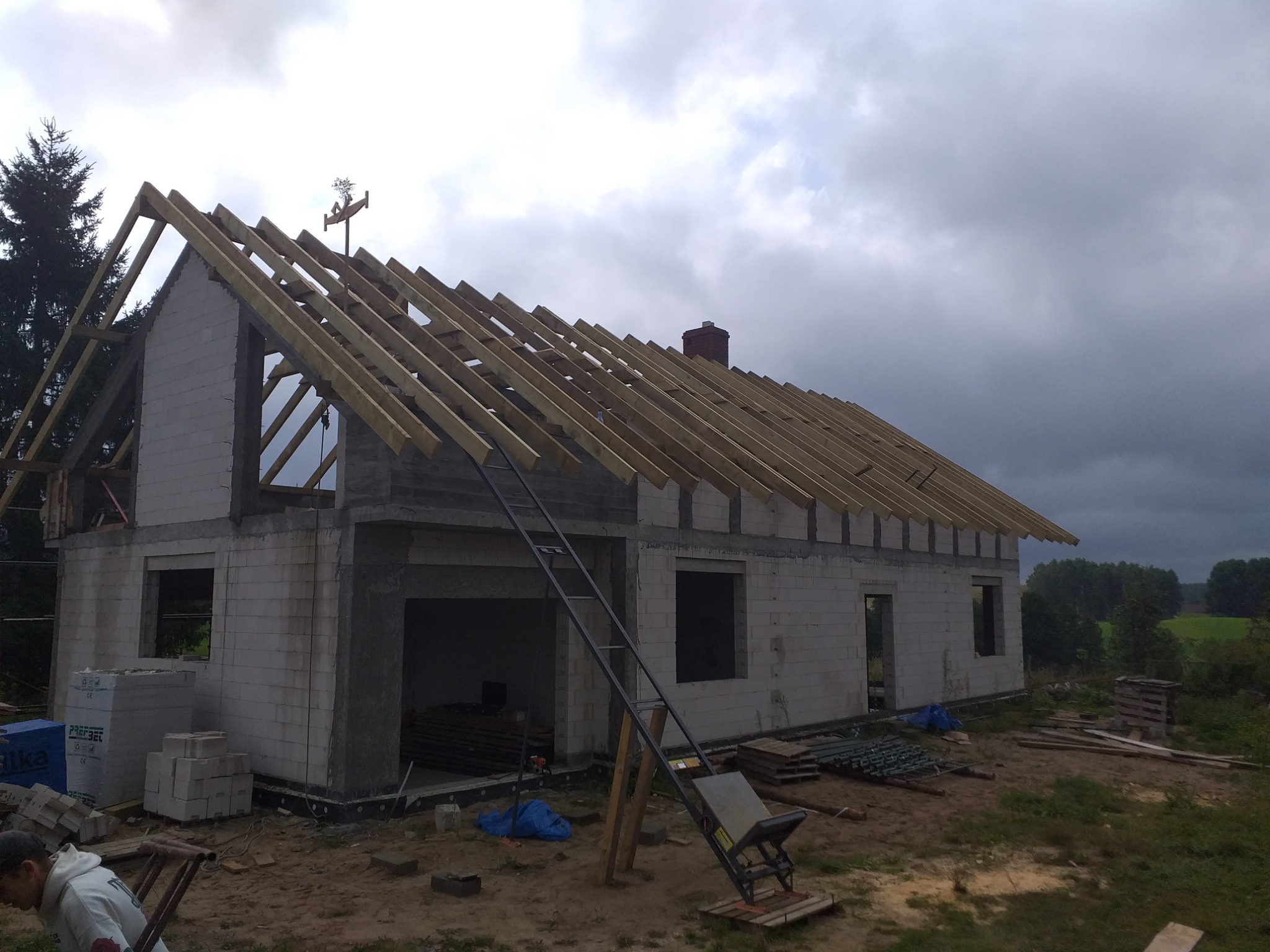 Surowy stan budynku z pustaków z drewnianą konstrukcją dachu w trakcie budowy, widoczne narzędzia i materiały budowlane.