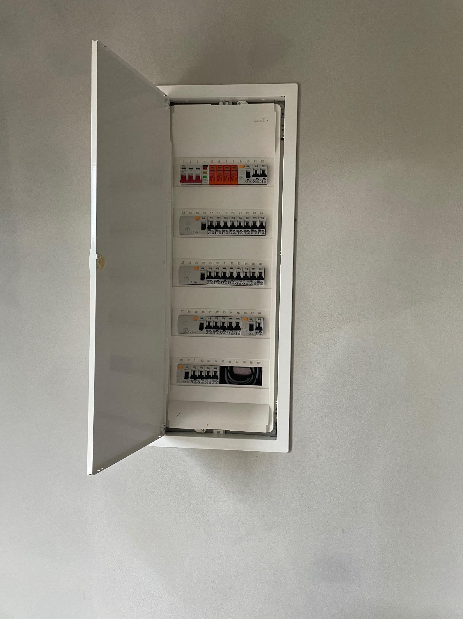 Otwarta skrzynka bezpiecznikowa z widocznymi bezpiecznikami i numeracją na szarej ścianie. Widok z bliska na zamontowaną rozdzielnicę elektryczną.